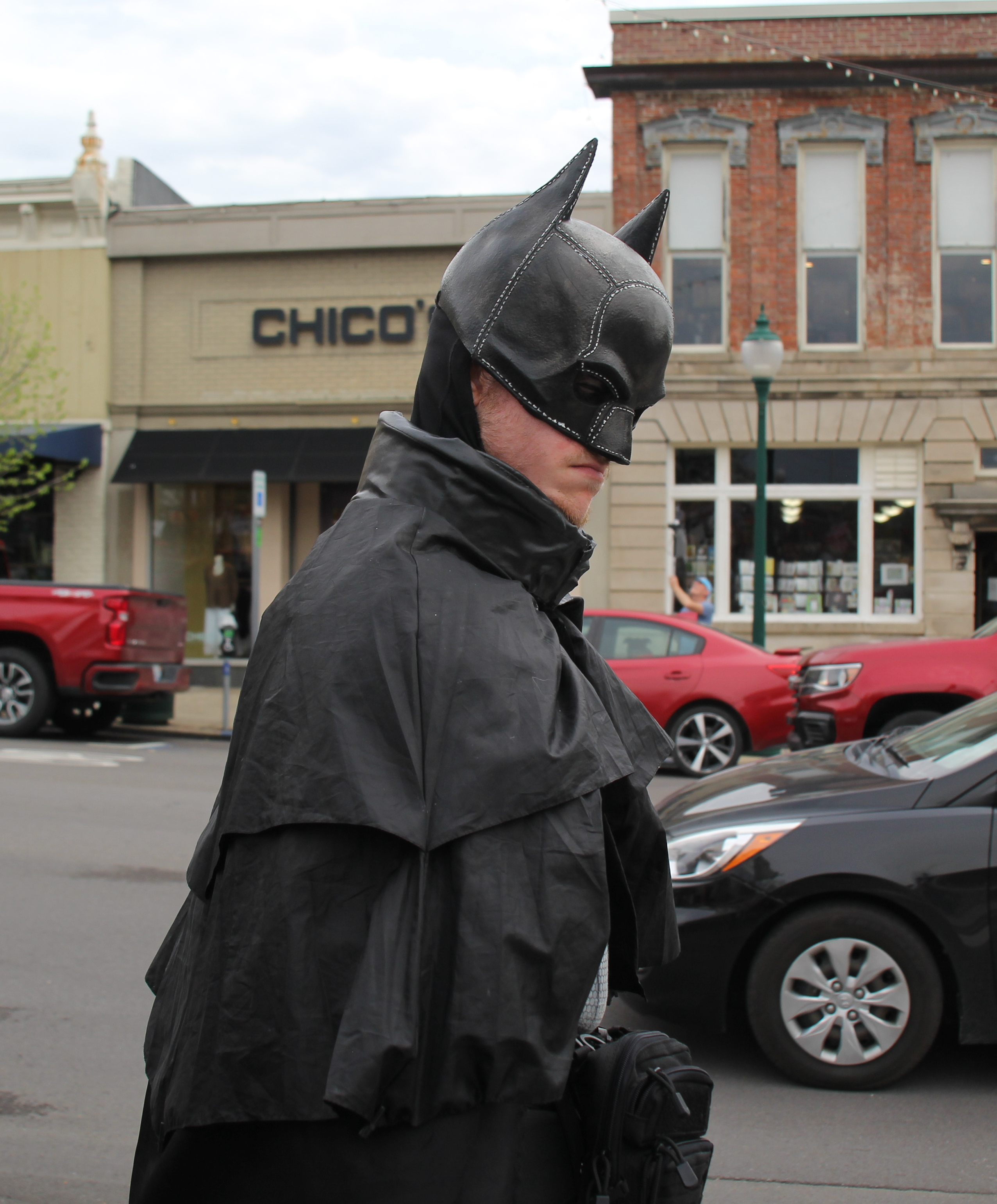 a man dressed in a bat man costume stands on a city street, looking back over his shoulder at the camera