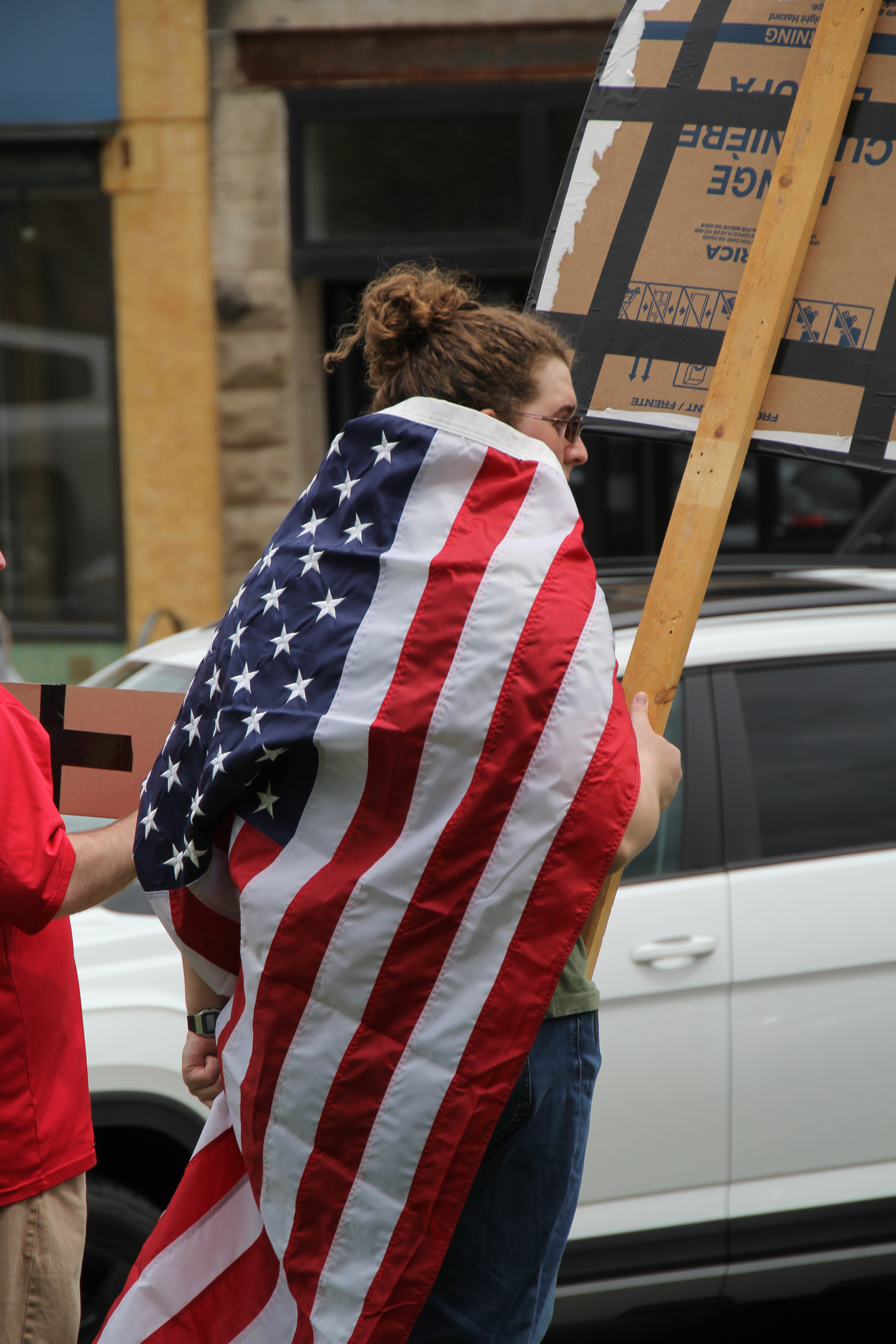 a woman using the american flag as a cape holds a sign with her back to the camera. she has red hair and sunglasses