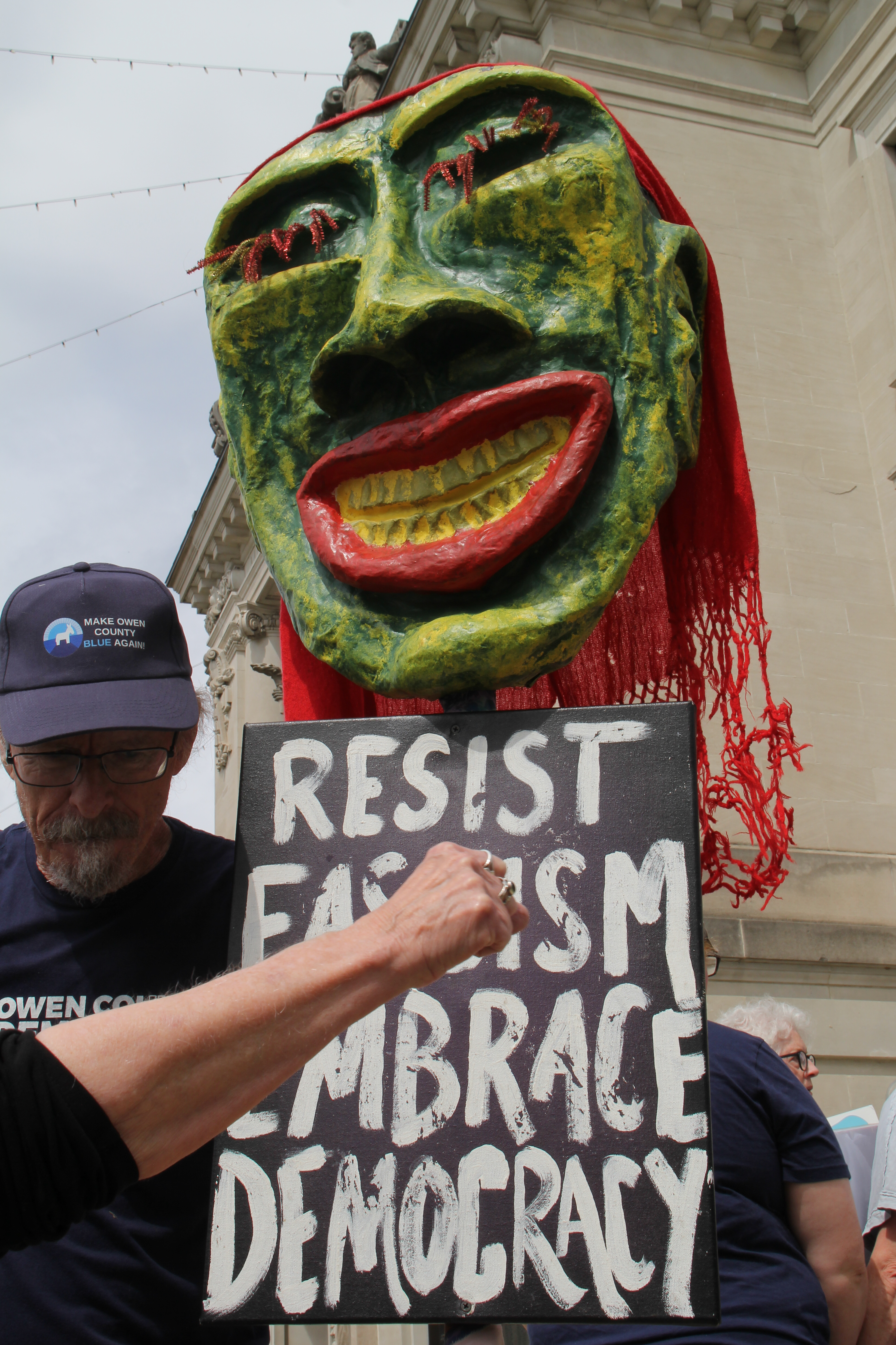 a giant green head paper-mache sculpture with red lips, red pipe cleaner eye lashes, and red cloth for hair sits on a sigh that says resist fascism, embrace democracy. a man with a hat stands to the left of it. a fist in the foreground strikes upward through the sign, making it look like the fist is punching through fascism
