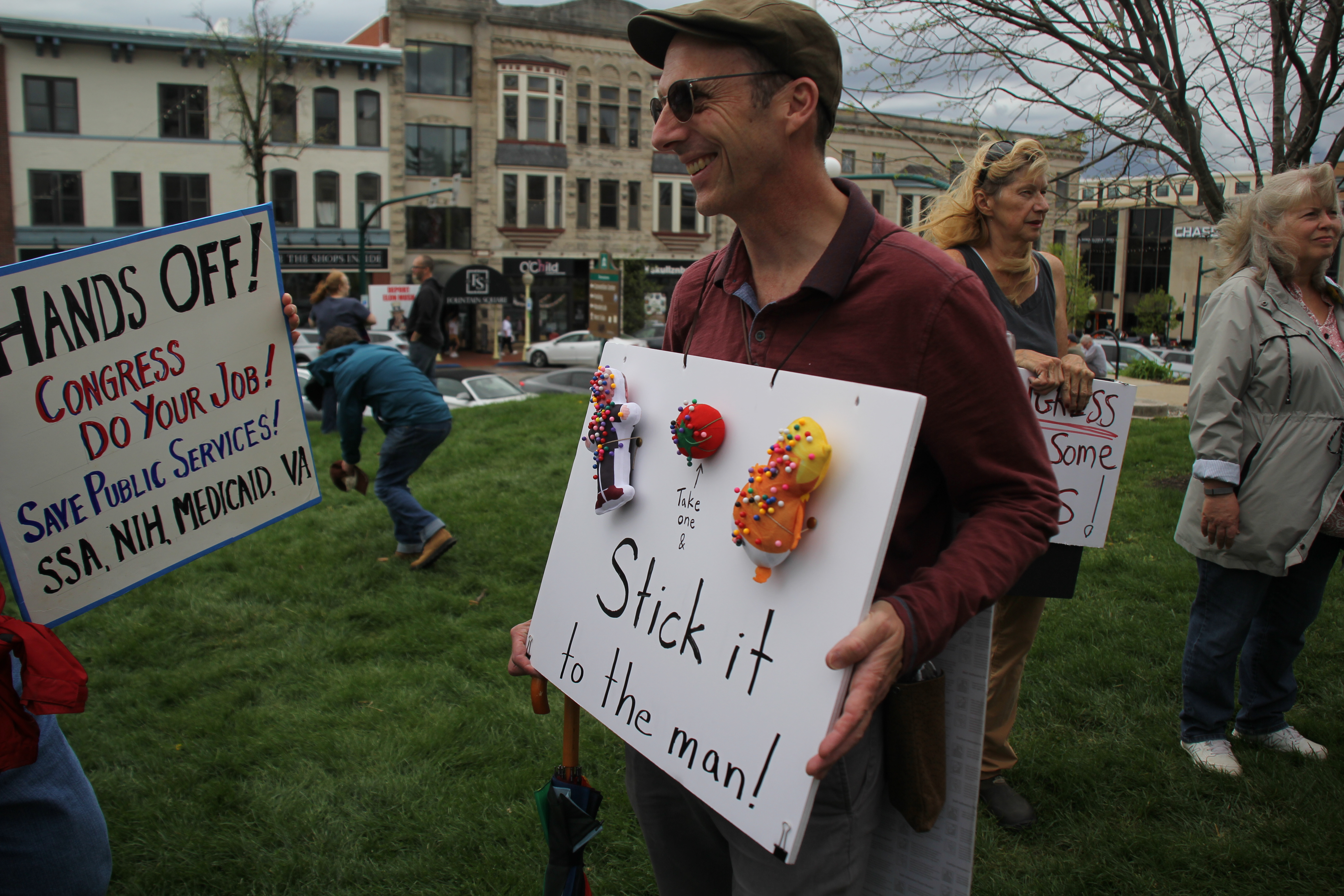 a man with a cap and sunglasses wears a sign around his neck. the sign says stick it to the man! a tomato pincushion is flanked by two donald trump themed pincushions.