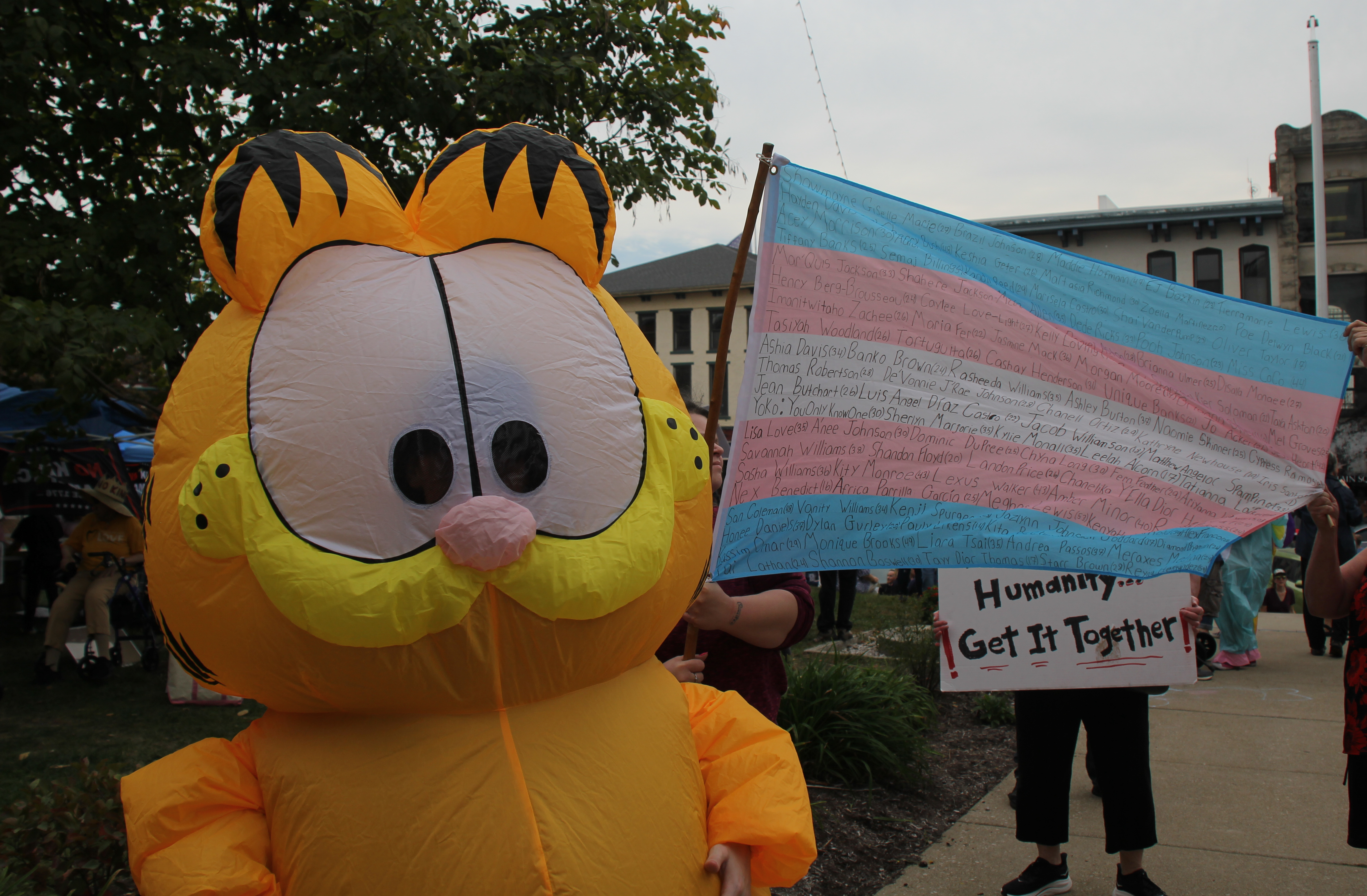 A person in a large inflatable Garfield costume stands next to a trans pride flag. Every inch of the flag has names of murdered trans people written on it.